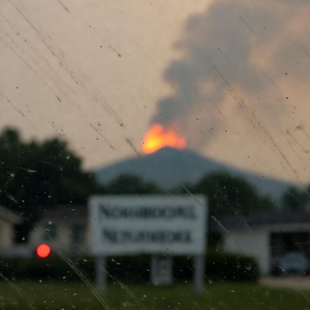 Blurry phone shot through dirty windshield: flames crest ridge behind neighborhood sign.